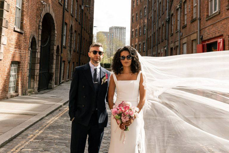 Nina & Dom stand in a industrial Manchester city centre alley way surrounded by red brick charm and holding stunning Wedding florals